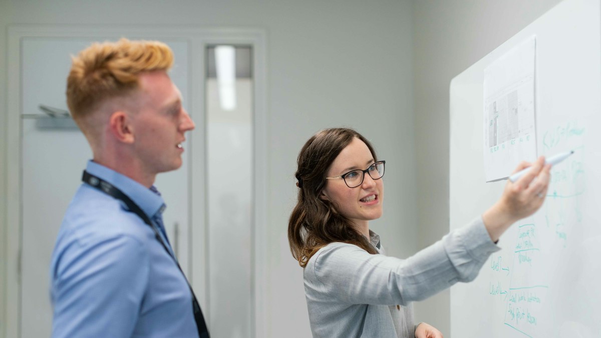 An engineer smiles as she points to a sketch on a whiteboard while a colleague looks on
