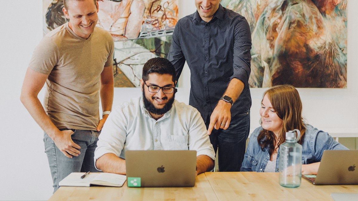 Four colleagues smile round a laptop at a table as one of them points at the screen