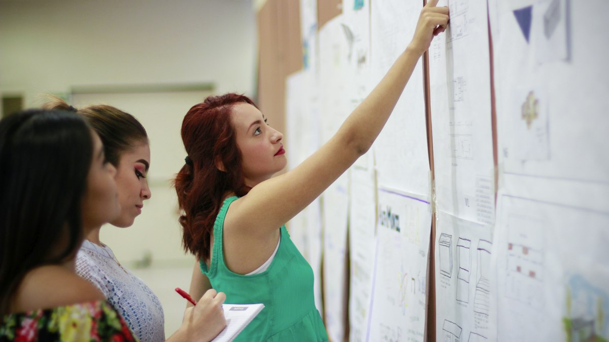Two women reading through papers pinned to a wall, one taking notes